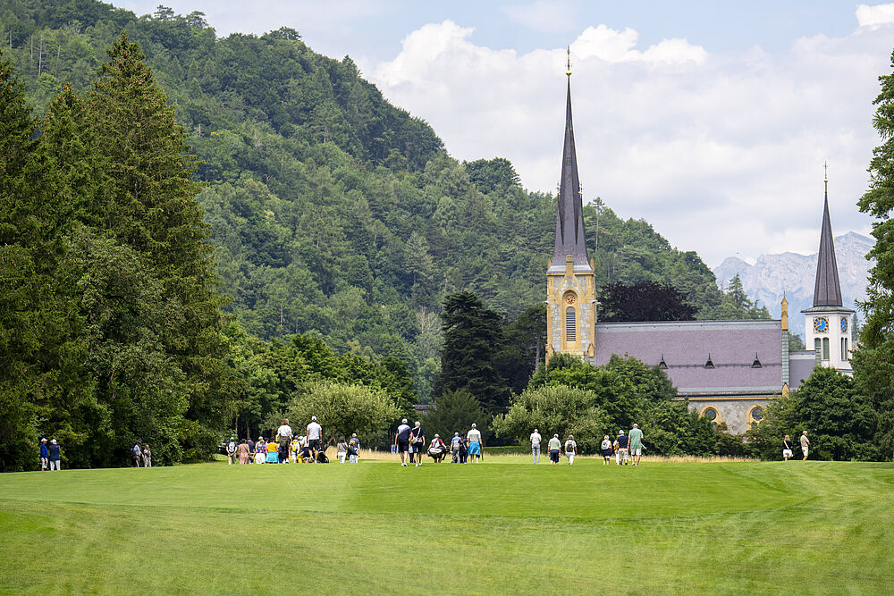 Ein malerischer Blick auf einen grasbewachsenen Park mit Menschen, die sich auf dem Golfplatz aufhalten. Eine Kirche mit hohen Türmen und üppig grünen Hügeln bildet einen ruhigen Hintergrund unter einem teilweise bewölkten Himmel.