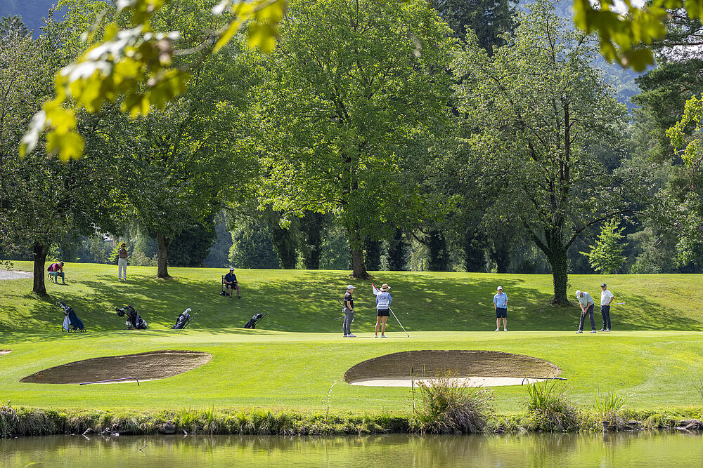Eine ruhige Parkszene mit spazierenden und sitzenden Menschen unter üppig grünen Bäumen. Das Sonnenlicht fällt durch die Blätter und schafft eine ruhige und friedliche Atmosphäre.