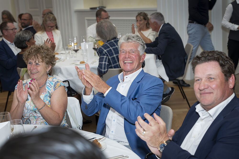 Drei Personen, die beim Gala Abend an einem runden Tisch mit weißem Tischtuch sitzen, klatschen und lächeln. Der Raum ist mit anderen Gästen gefüllt, die die Veranstaltung genießen.