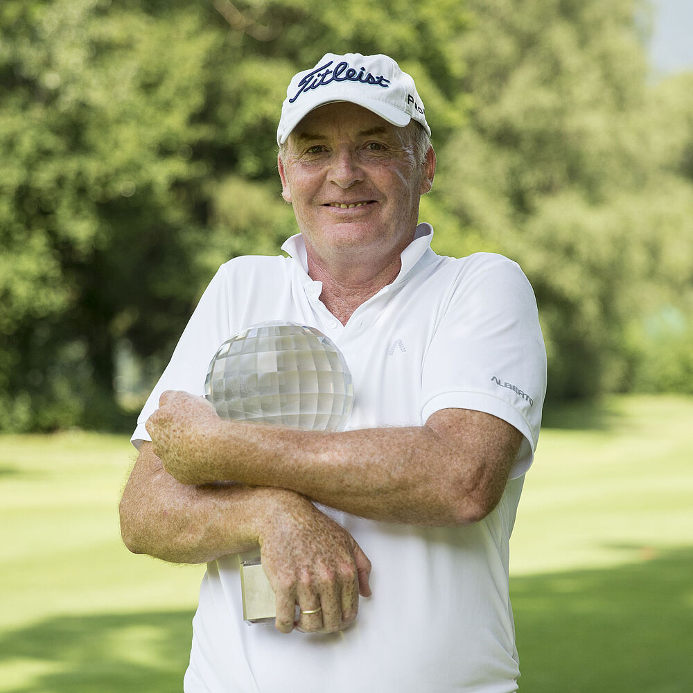 Ein lächelnder Gordon Manson in weißem Golfhemd und Mütze steht auf dem Golfplatz und hält eine Trophäe in den Armen. Die Stimmung ist fröhlich und entspannt.
