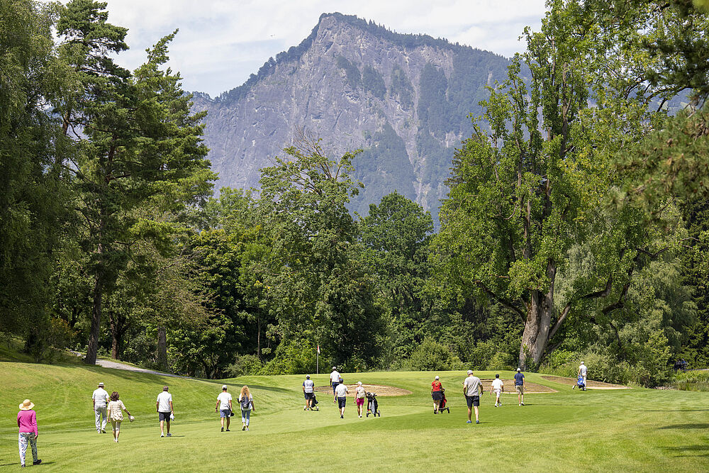 Eine Gruppe von Menschen läuft über den Golfplatz, die von großen Bäumen umgeben ist, mit einem Berg im Hintergrund unter einem teilweise bewölkten Himmel. Die Szene ist lebhaft und fröhlich.