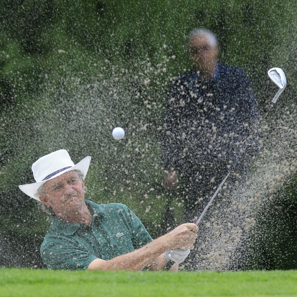 Davis Creamer mit weißem Hut und grünem Hemd schlägt einen Golfball aus einem Bunker ab, wobei Sand umherfliegt. Eine zweite Person schaut im üppig grünen Hintergrund zu.