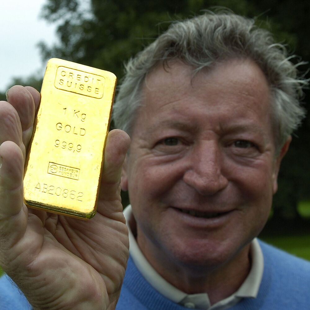 Der lächelnde Maurice Bembridge hält im Freien stehend einen Goldbarren mit eingraviertem Text. Der Hintergrund ist eine verschwommene Ansicht von grünem Laub, die ein Gefühl von Erfolg vermittelt.