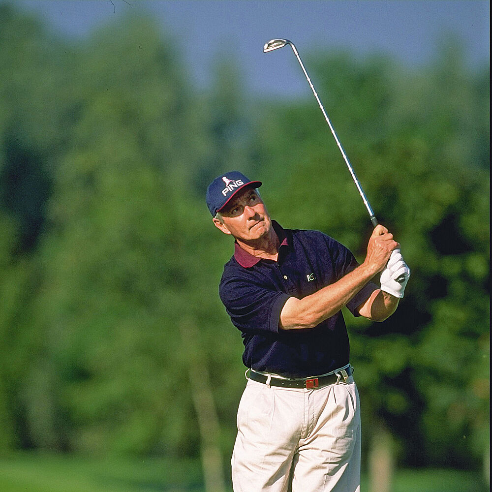 Brian Wates in einem marineblauen Hemd und weißen Handschuhen schwingt einen Schläger auf einem üppig grünen Golfplatz. Bäume und ein klarer blauer Himmel bilden die ruhige, konzentrierte Kulisse.