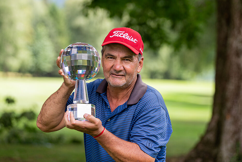 José Coceres in roter Mütze und blauem Hemd lächelt und hält den glänzenden Pokal in die Höhe. Im Hintergrund sind grüne Bäume und Gras zu sehen, was eine feierliche Stimmung vermittelt.