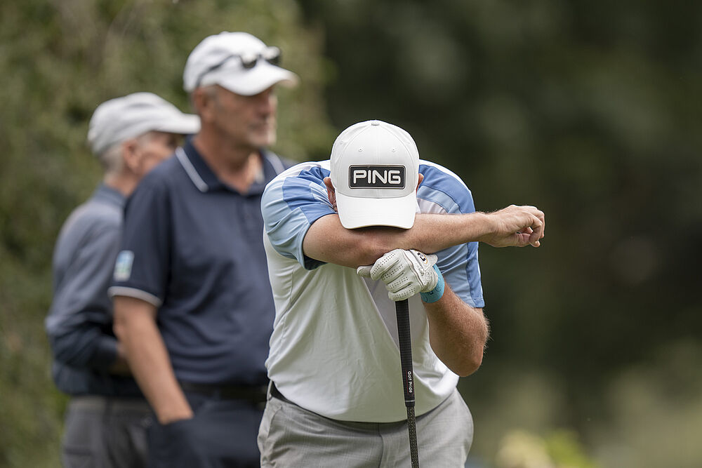 Ein Golfer mit einer hellen Mütze und einem hellen Hemd stützt sich frustriert auf seinen Schläger. Zwei weitere Männer mit Hüten stehen im verschwommenen grünen Hintergrund.