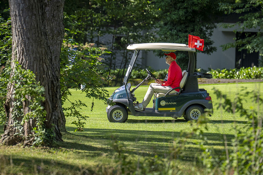 Eine Person in einem roten Hemd fährt einen Golfwagen auf einem sonnenbeschienenen, grasbewachsenen Rasen. Im Hintergrund sind ein großer Baum und üppiges Grün zu sehen.