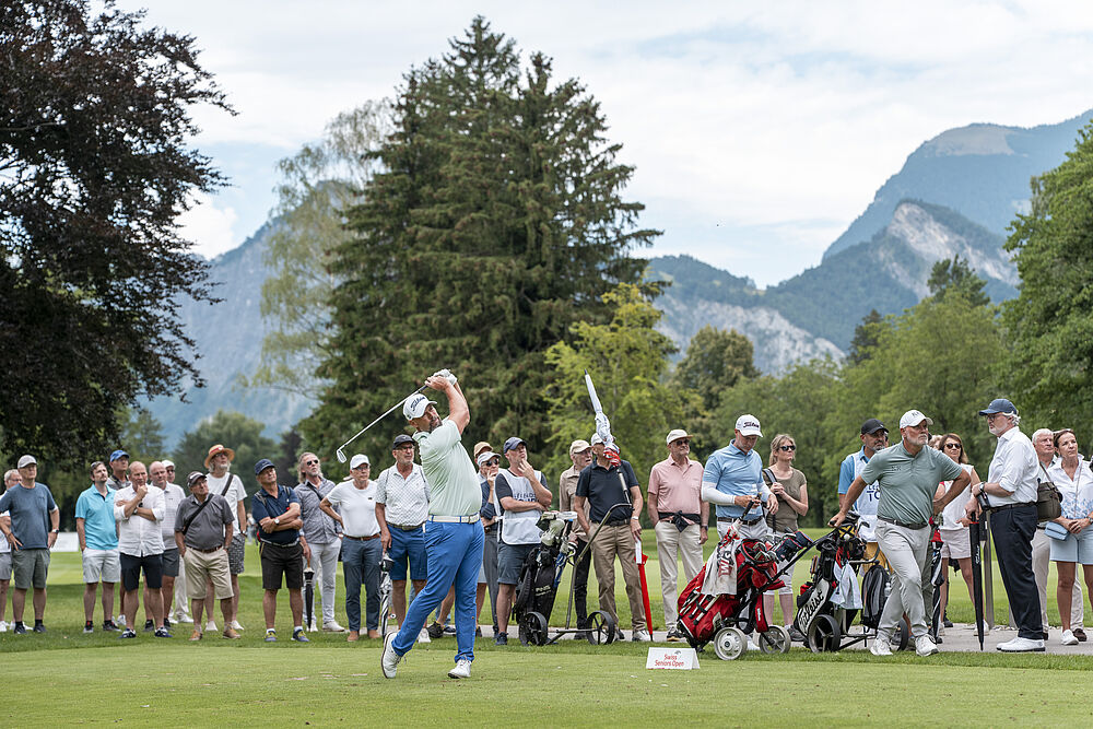 Ein Golfer in blauer Hose schwingt den Schläger auf einem üppigen grünen Platz, umgeben von einer großen Menschenmenge und vor einer Kulisse aus Bäumen und Bergen.