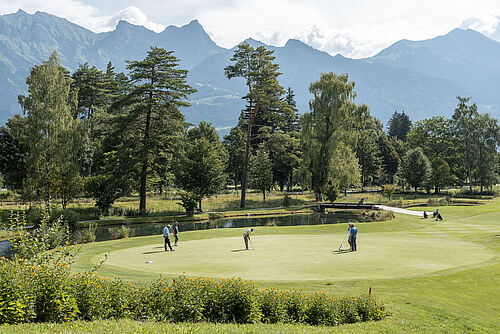 Ein ruhiger Golfplatz mit drei Golfern auf einem Grün, umgeben von lebhaften Bäumen und fernen Bergen unter einem bewölkten Himmel, der Ruhe vermittelt.