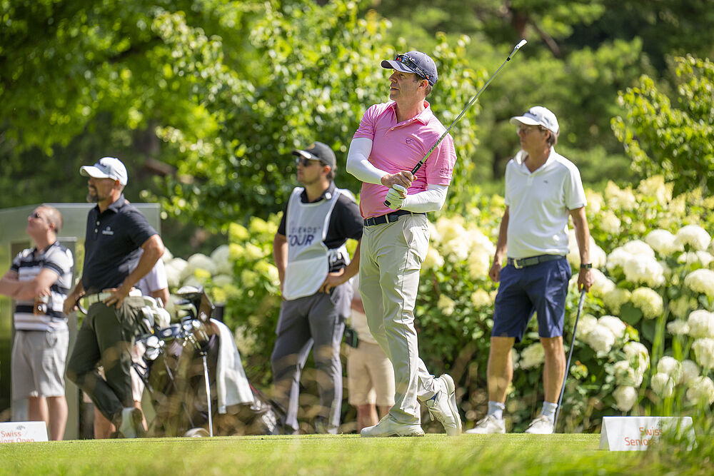 Ein Golfer in einem rosa gemusterten Hemd schwingt an einem sonnigen Tag den Schläger, umgeben von grünen Bäumen und drei Zuschauern, die eine entspannte und konzentrierte Stimmung vermitteln.