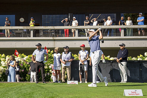 André Bossert in blauem Hemd und weißer Hose schwingt seinen Golfschläger bei Abschlag 1, beobachtet von einer Menge von Zuschauern und Kindern, mit Blumen im Hintergrund.