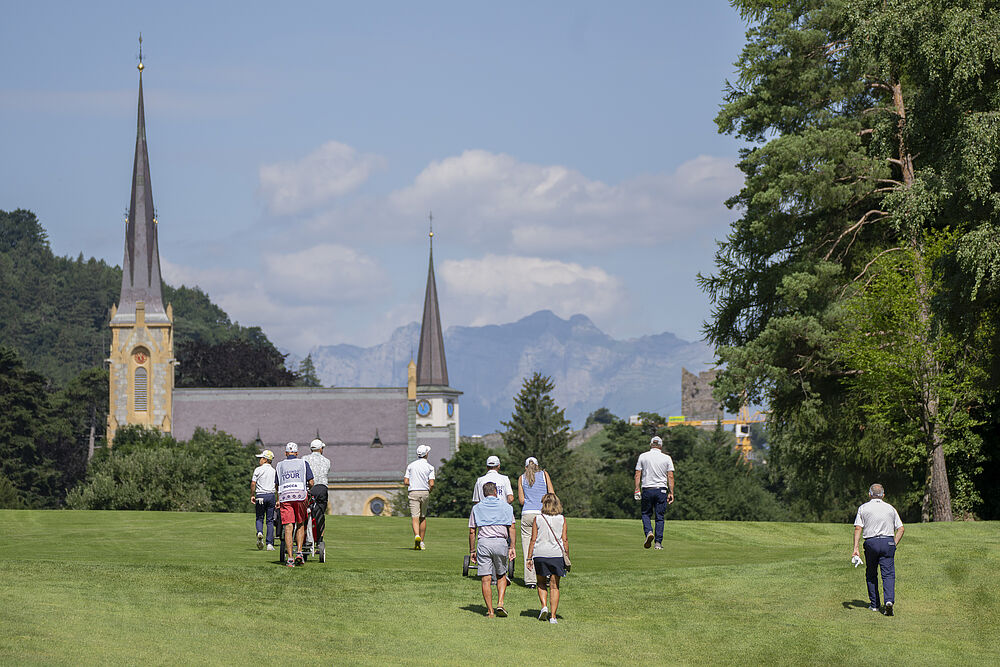 Eine Gruppe von Menschen geht über einen Golfplatz mit einer Kirche und Bergen im Hintergrund. Die Szene ist sonnig, mit einer entspannten und fröhlichen Atmosphäre.