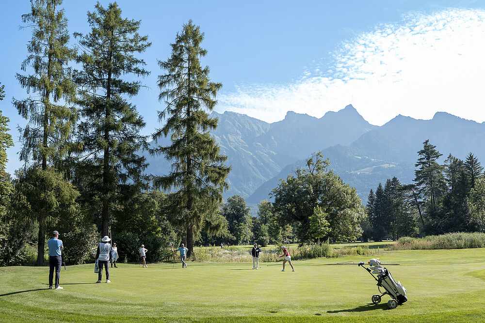 Golfer spielen auf einem grünen Platz mit hohen Bäumen und Bergen im Hintergrund. Der Himmel ist klar mit vereinzelten Wolken, die eine heitere Atmosphäre schaffen.
