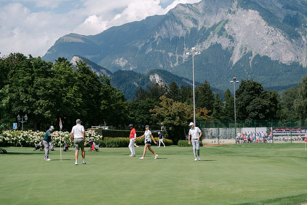 Eine Gruppe von Golfspielern auf einem üppigen grünen Platz mit dramatischen Bergen und einem teilweise bewölkten Himmel im Hintergrund, der eine ruhige Szene schafft.