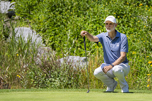 André Bossert in der Hocke auf dem Green. Er stütz sich am Putter und trägt ein blaues Shirt mit weissem Cap. Im Hintergrund ist viel grün und er wirkt überlegt. 