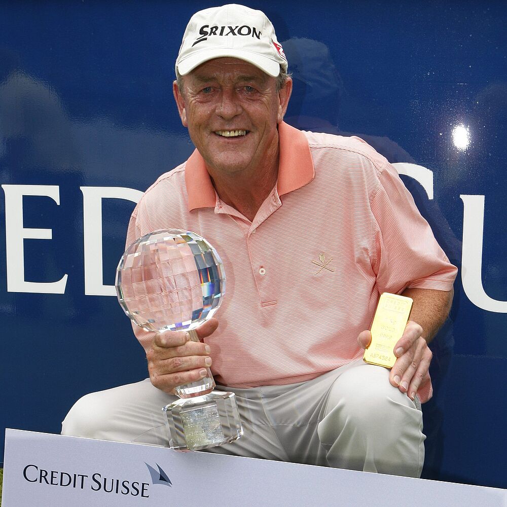 Carl Mason im rosa Hemd und weißer Mütze lächelt und hält eine Golftrophäe in der Hand. Er hockt neben einem Schild der Credit Suisse, das Stolz und Erfolg ausdrückt.