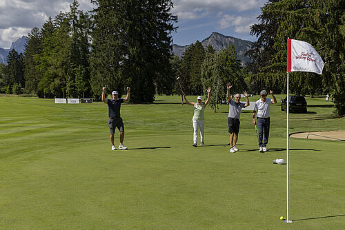 Vier Personen feiern mit erhobenen Armen auf einem sonnigen Golfplatz neben einer Flagge. Im Hintergrund sind üppige Bäume und Berge zu sehen. Die Stimmung ist fröhlich.