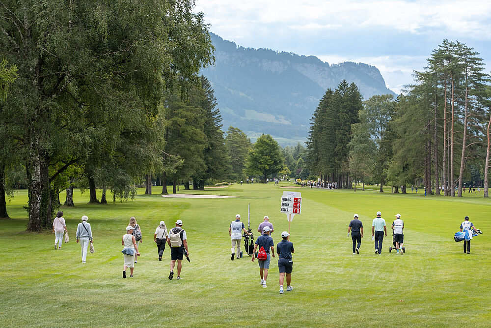 Zuschauer folgen den Golfern auf dem Golfplatz. Im Hintergrund befinden sich Berge und links und rechts sind Bäume.
